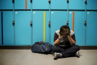 Child sitting on the ground, hands on face upset in a school hallway.