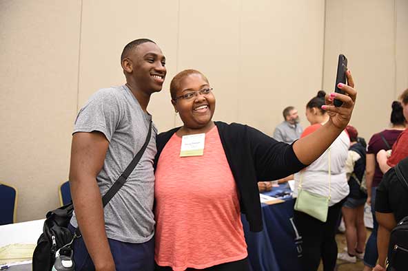 Two people standing next to each other posing for a picture using a cellphone