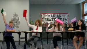 Group of students in a classroom sitting at desks raising their hands