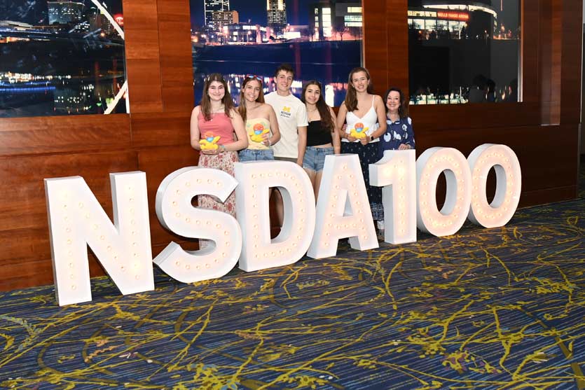 Students and their coach at the National Tournament standing behind the NSDA 100 Sign